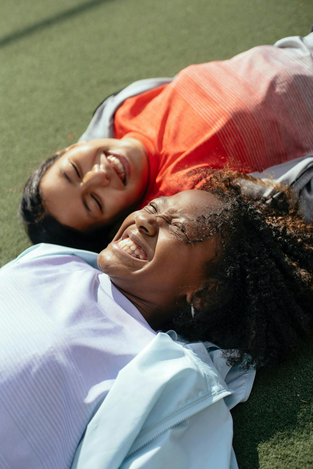 Two individuals are lying on a green outdoor surface, smiling and enjoying the sun while wearing light-colored clothing.