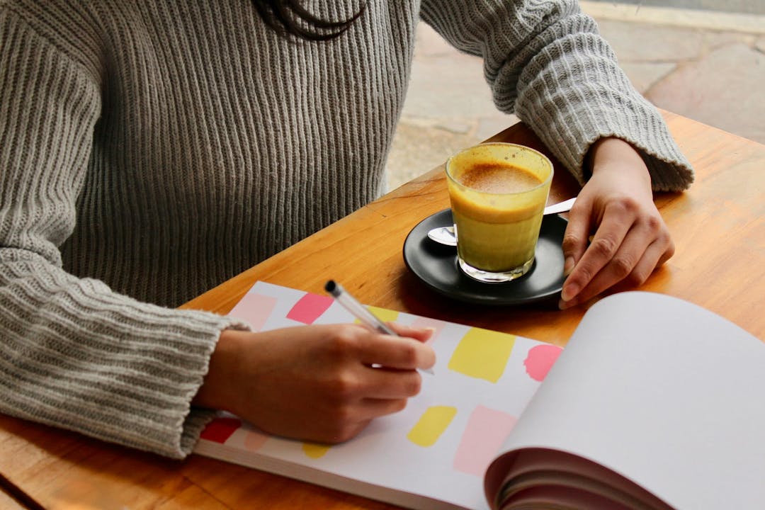 A person writes in a colorful notebook while sitting at a wooden table, with a glass of coffee on a black saucer nearby.