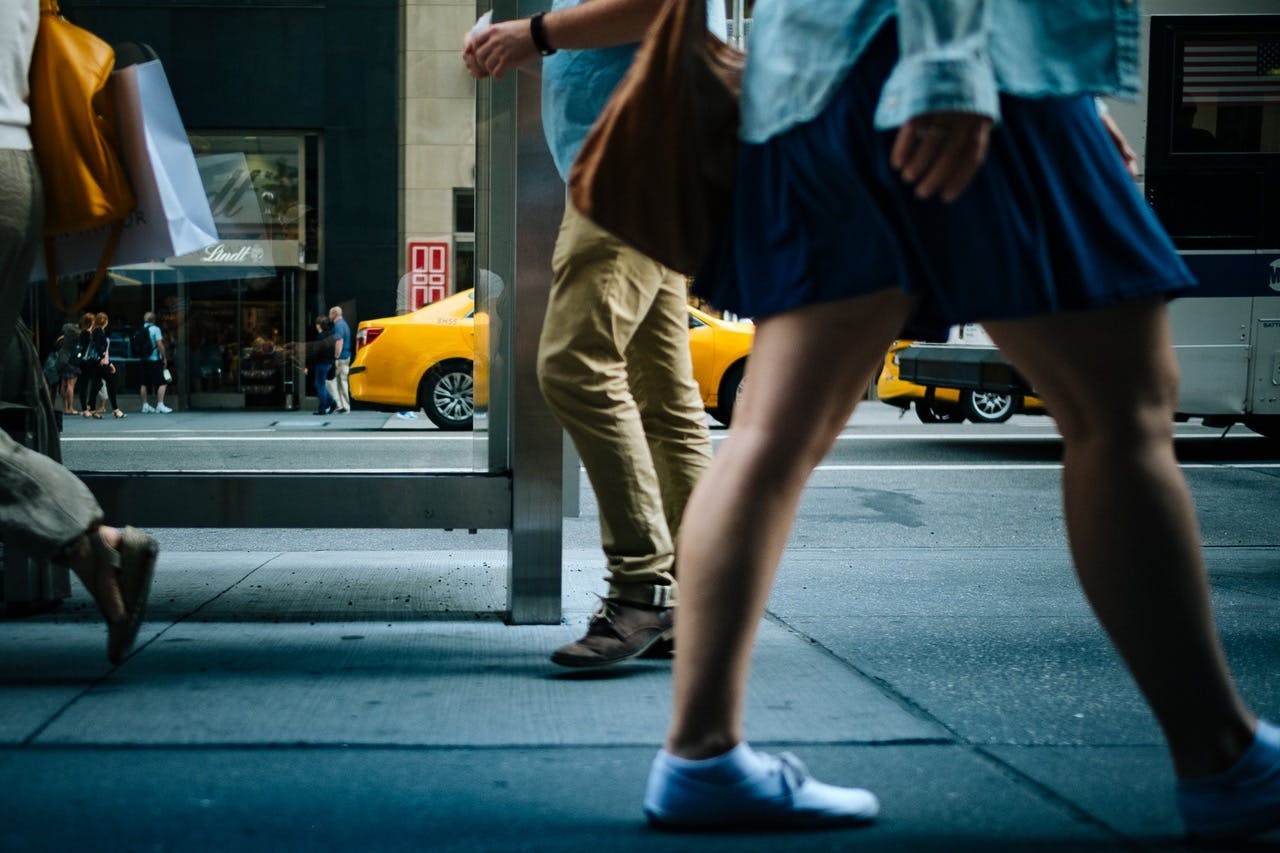 Pedestrians walking on a city sidewalk with yellow taxis driving by in the background, near storefronts including a Lindt shop. People's midsections and legs are primarily visible.