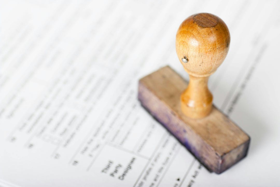 A wooden rubber stamp rests on a stack of documents with text, including "Third Party Designee," "Sign," and "Date."