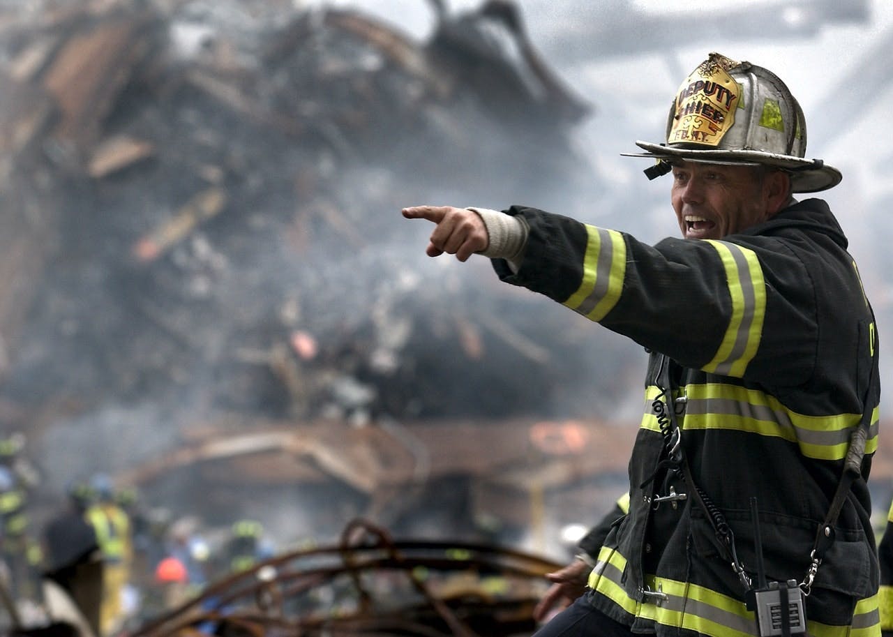 Firefighter in protective gear points urgently amidst smoky debris and rescue workers in helmets and reflective vests, possibly directing operations at a disaster site. "DEPUTY CHIEF" text visible on helmet.