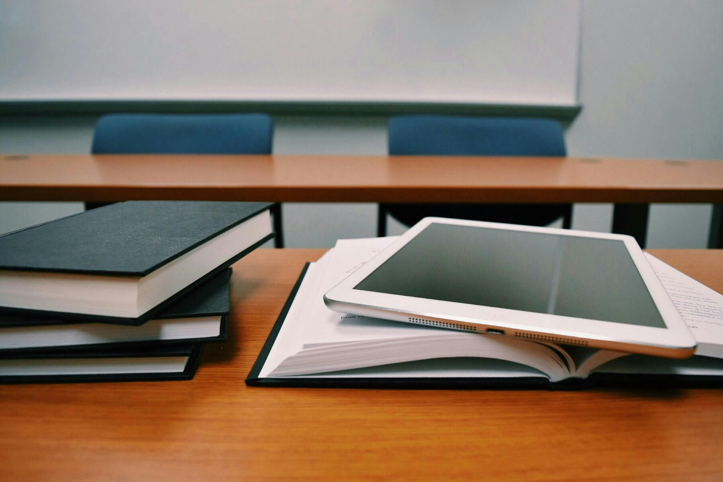 A tablet rests on an open book next to a stack of closed books on a wooden desk in a classroom with blue chairs and a whiteboard in the background.