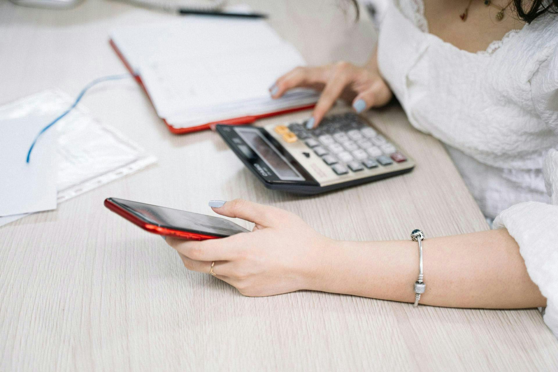 A person uses a calculator with one hand while holding a smartphone in the other, at a desk with papers and an open notebook.