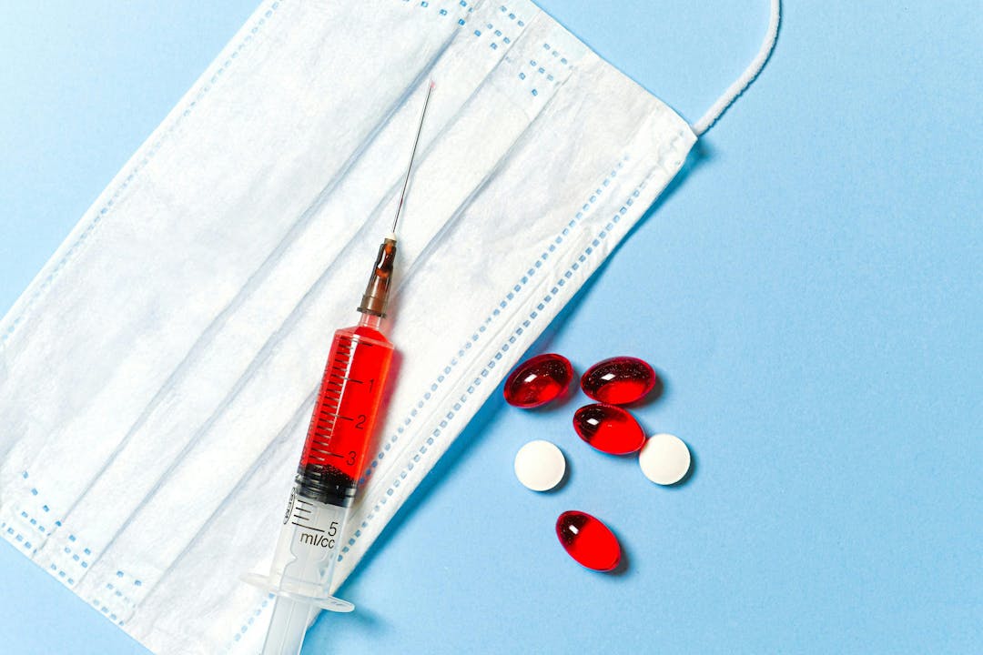 Syringe filled with red liquid placed on a white face mask, accompanied by red oval pills and round white tablets, on a light blue background.