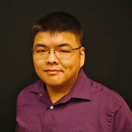 A man wearing glasses and a purple button-up shirt smiles slightly, standing against a plain black background.