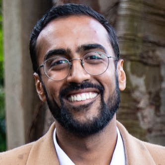 A smiling man wearing glasses and a tan coat stands outdoors, with a pillar and trees in the background.