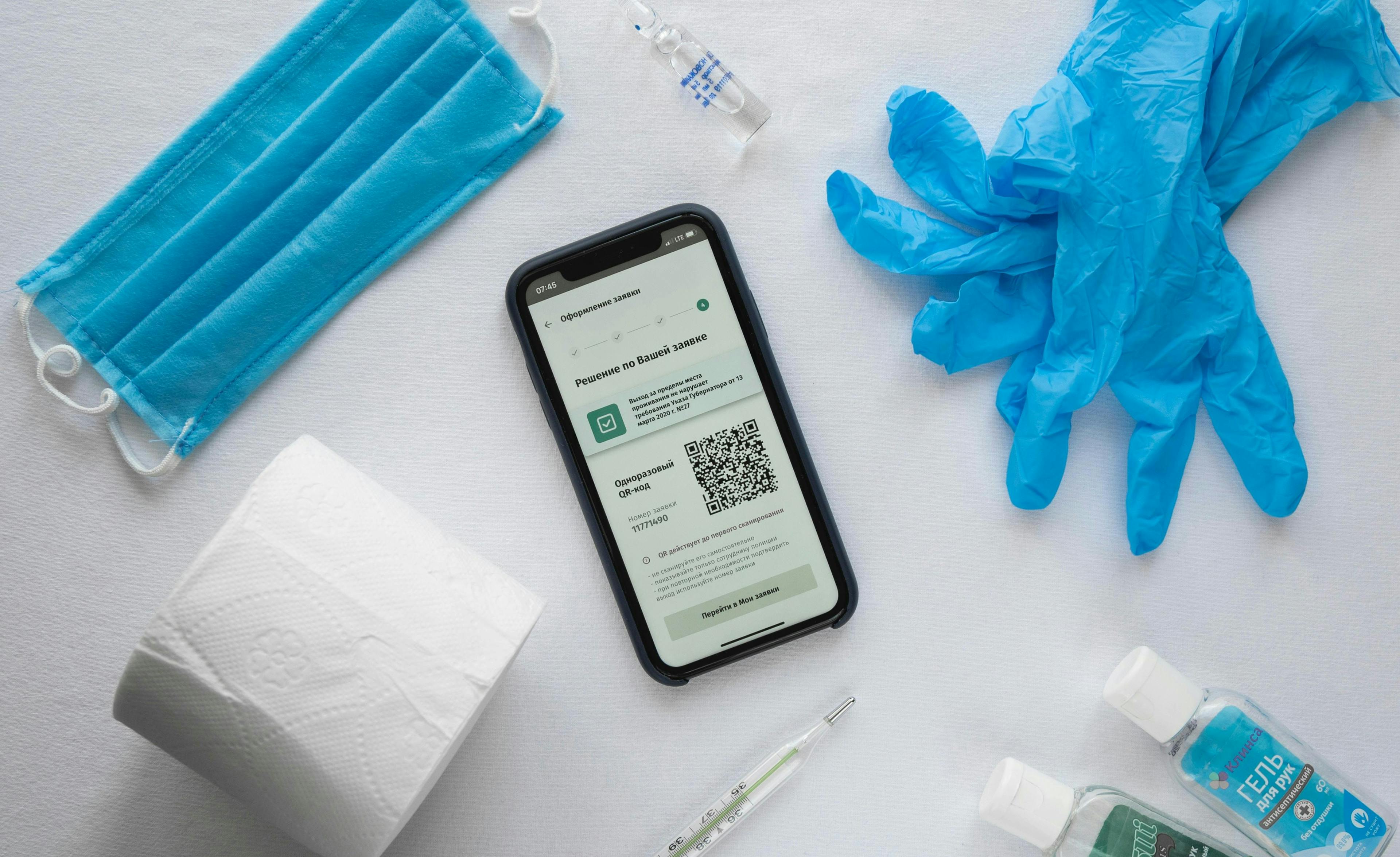 A smartphone displays a QR code among a blue face mask, blue gloves, a roll of toilet paper, medical ampoule, thermometer, and hand sanitizers on a white surface.
