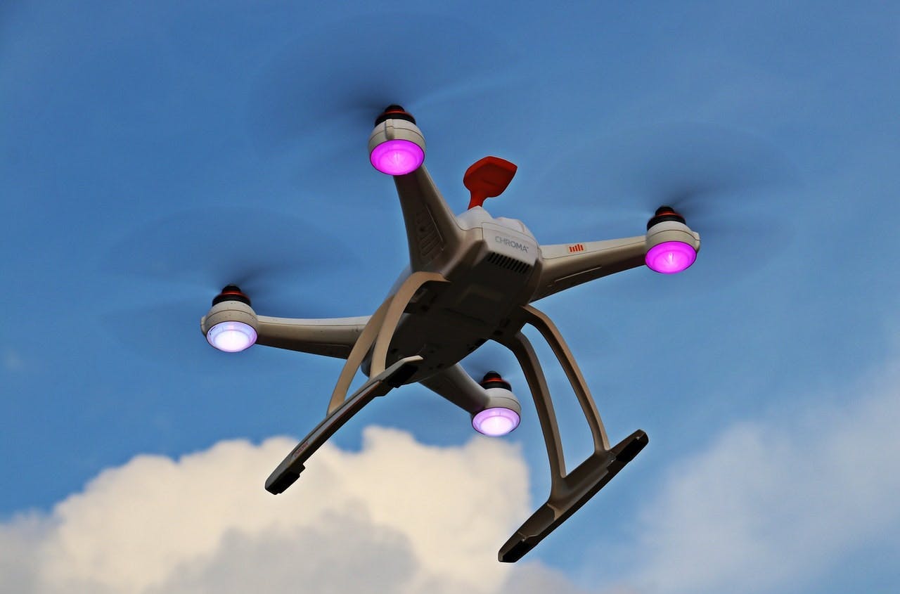 Quadcopter drone flying, equipped with purple-lit LED lights on each arm, set against a clear blue sky with few clouds. The drone has the word "dronar" inscribed on it.