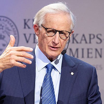An elderly man in a suit and tie speaks into a headset microphone, gesturing with his right hand, standing against a backdrop with partially visible text "KAPS," suggesting a formal event.
