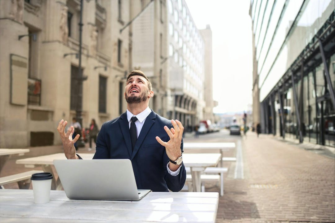 A man in a suit sits at an outdoor table with a laptop, raising his hands in frustration. He is surrounded by tall buildings on a deserted street.