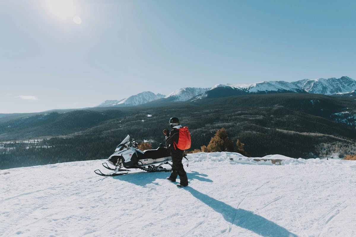 A man standing by a Skidoo, on top of a hill overlooking a valley. It’s winter.