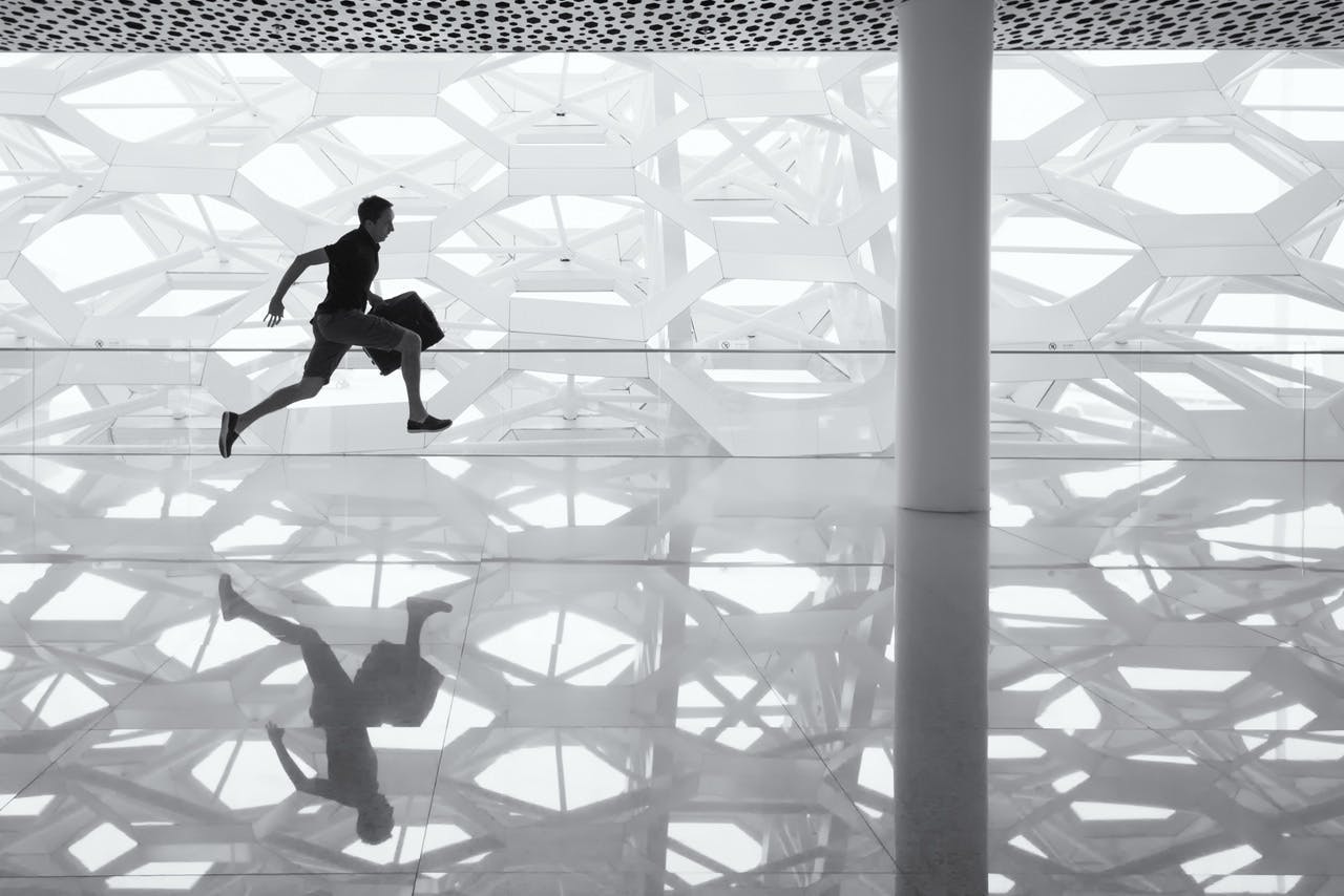 Person jumping, reflected on a shiny floor, indoors with hexagonal-patterned walls and ceiling, beside a pillar.