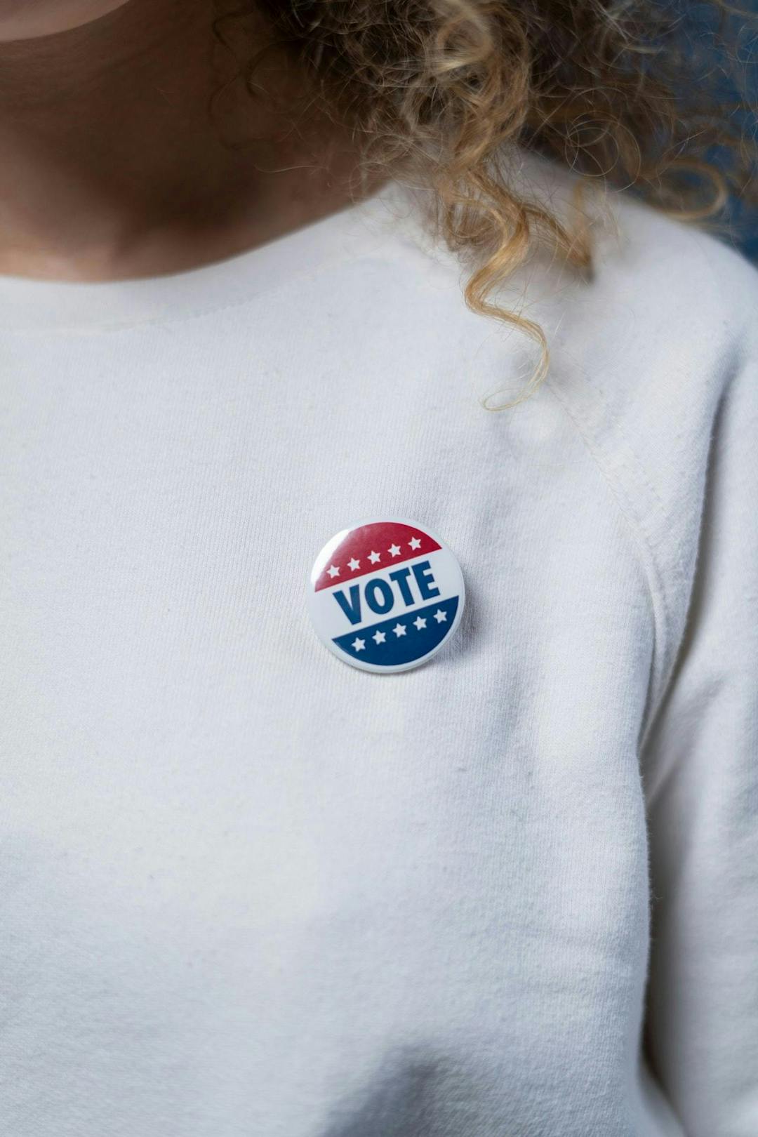 A person wearing a white shirt with a circular badge containing the word "VOTE" and stars in red, white, and blue.