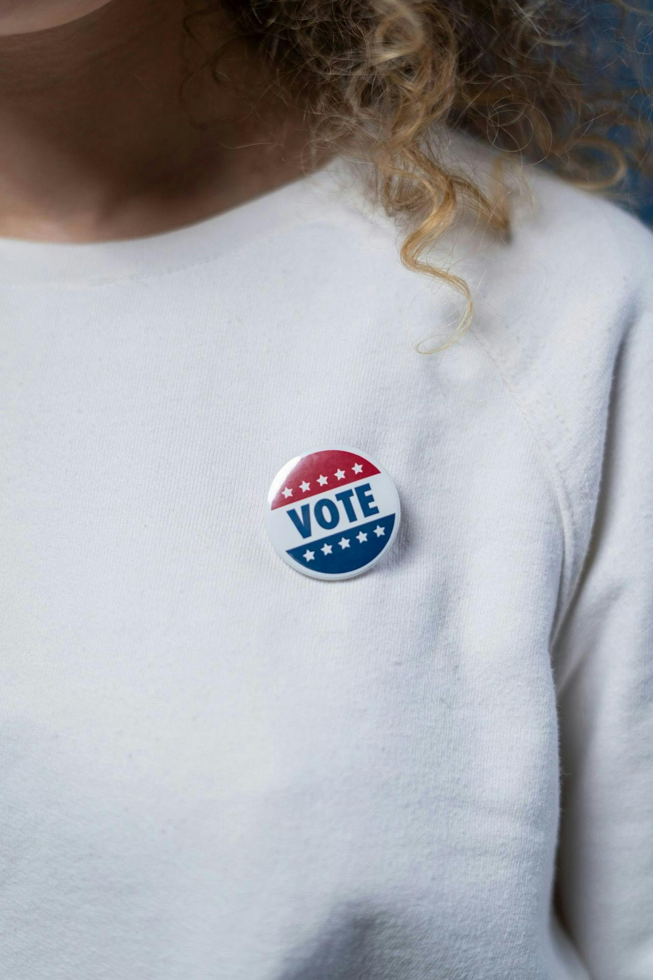 A person wearing a white shirt with a circular badge containing the word "VOTE" and stars in red, white, and blue.
