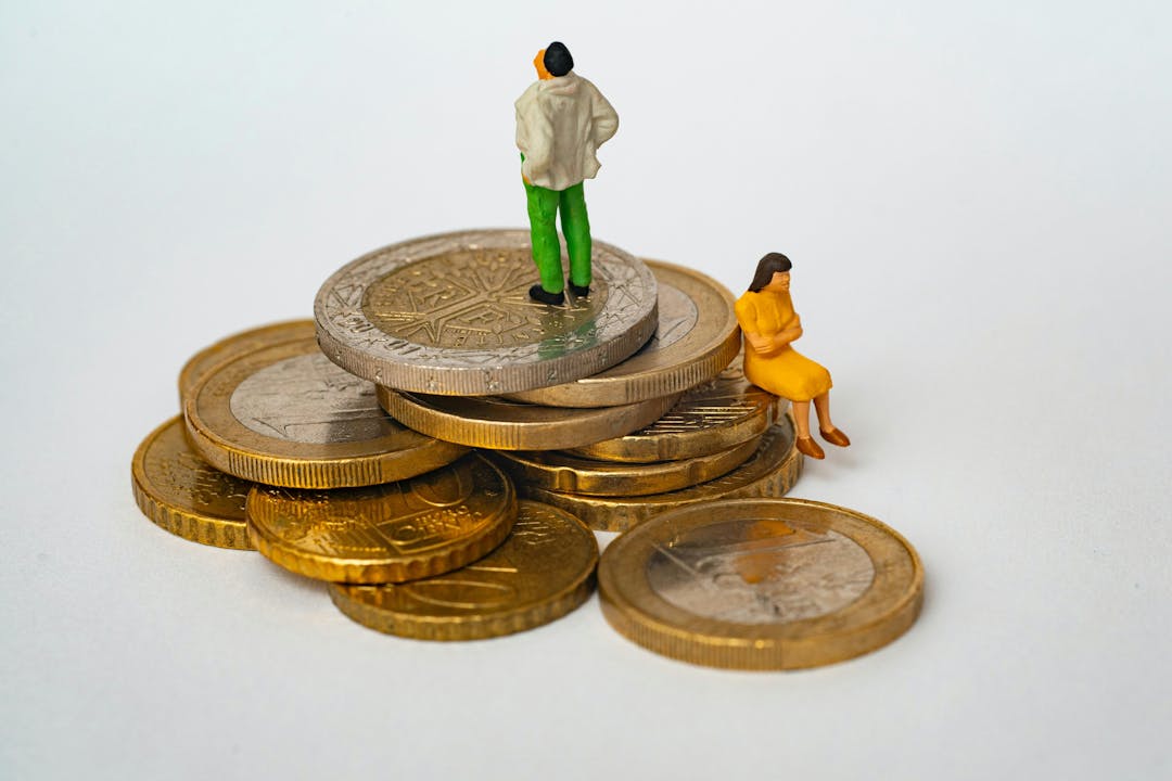 Miniature figures, one standing and one sitting, atop a stack of assorted coins in a white, featureless background. The standing figure gazes away, while the seated figure appears contemplative.