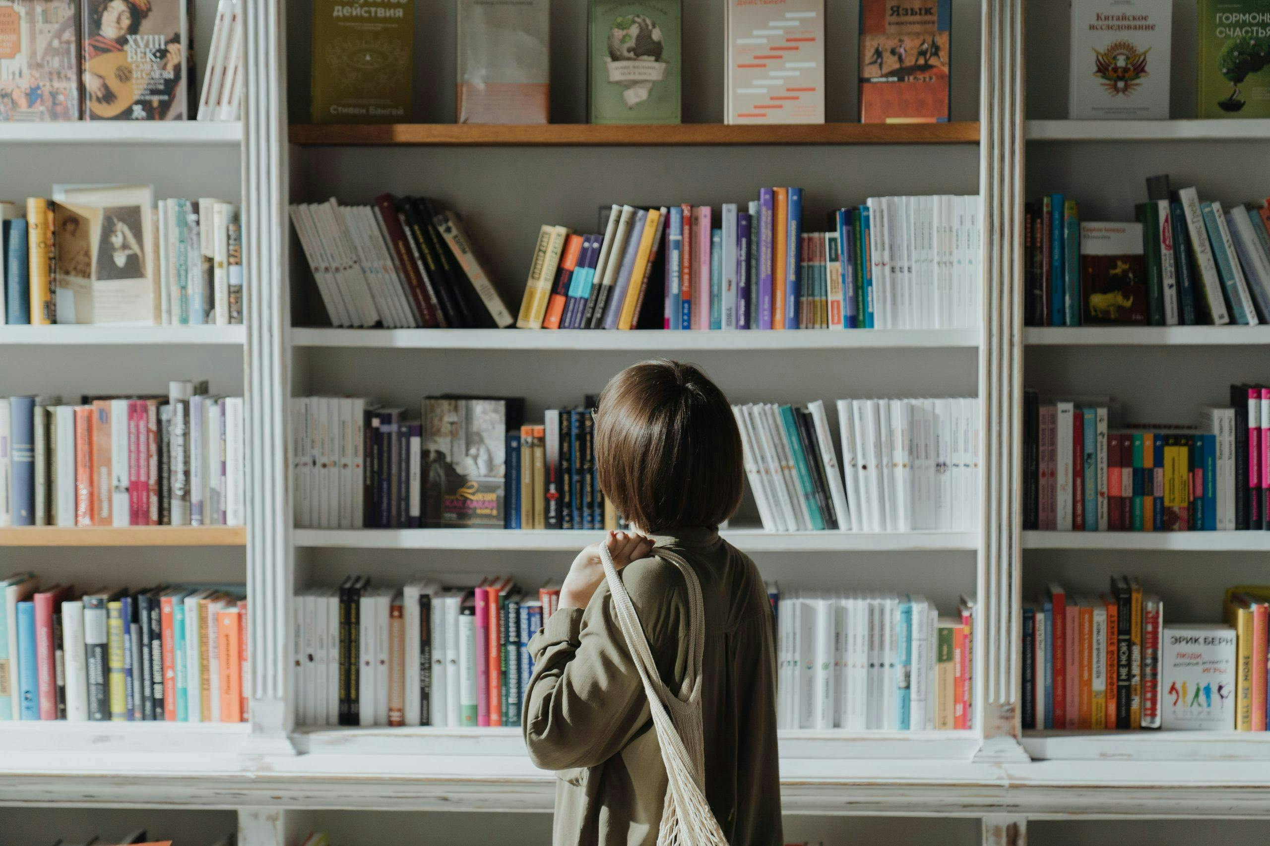 Person standing, browsing a large white bookshelf filled with various books, in a library or bookstore setting. They carry a beige tote bag and wear a green jacket.