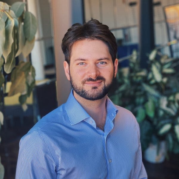 A man in a blue, striped shirt smiles while standing indoors, surrounded by green plants and modern office decor.