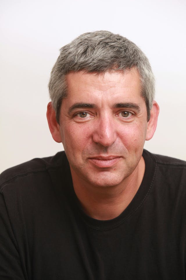 A man with short gray hair is smiling subtly, wearing a black shirt, and looking directly at the camera against a plain white background.
