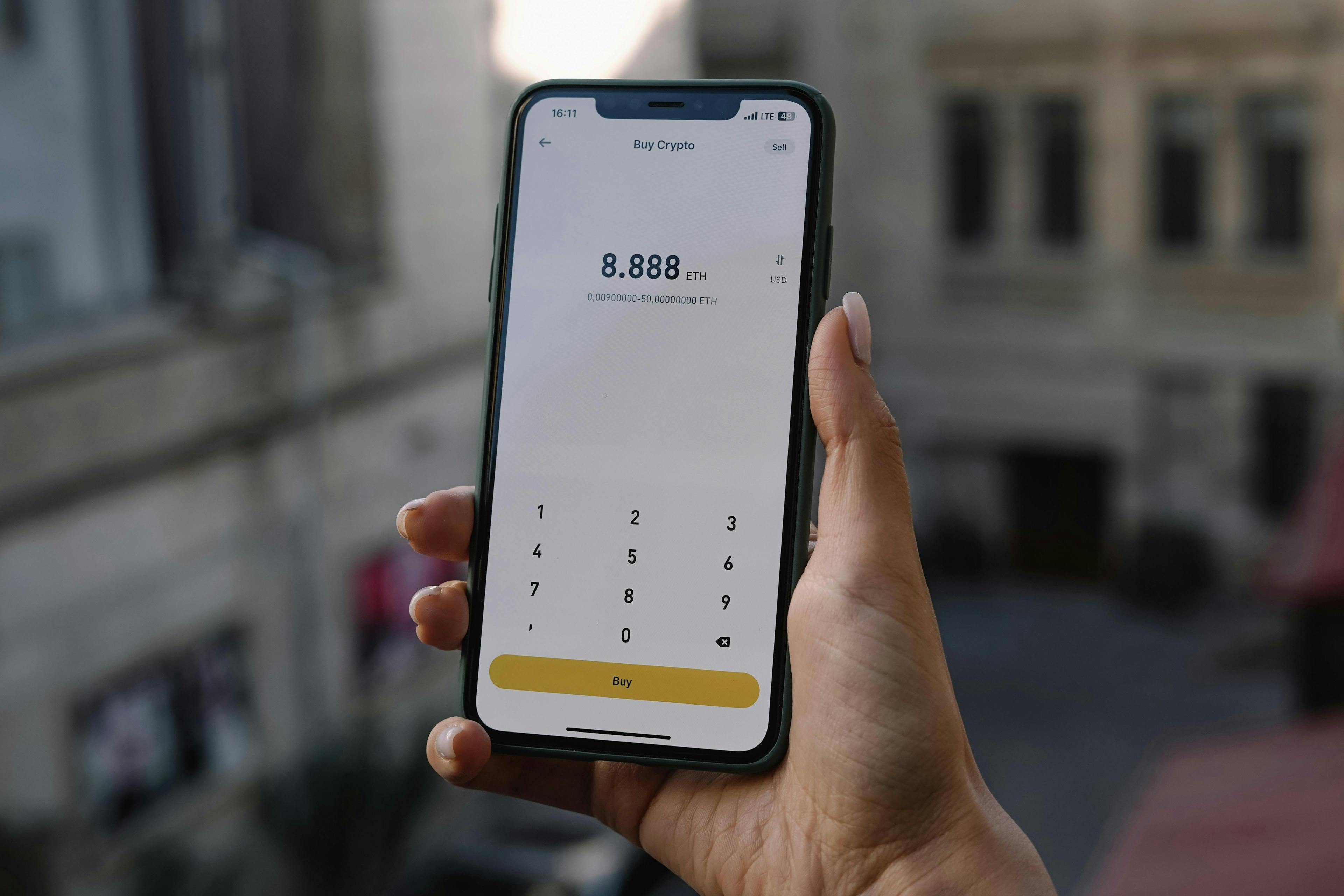 A hand holds a smartphone displaying a cryptocurrency purchase screen showing 8.888 ETH, with "Buy" and numeric keypad below. Background shows blurred urban buildings.