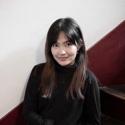 A young woman sits against a white and red wall, smiling slightly. She wears a black sweater, with straight hair falling over her shoulders, posed near some wooden stairs.