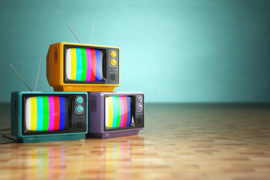 Three vintage televisions with rainbow screens, stacked in a pyramid arrangement, rest on a wooden floor against a turquoise wall.