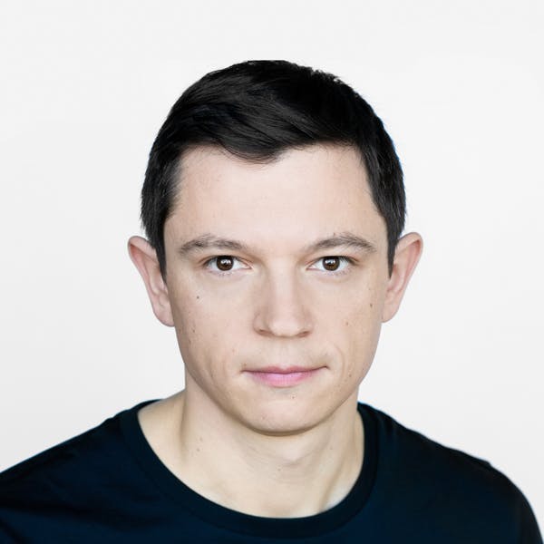 A person with short black hair looks directly into the camera, wearing a black shirt against a plain white background.
