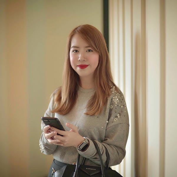 A woman with long hair holds a smartphone and wears a grey sweater adorned with beads, standing indoors near a beige textured wall, carrying a black handbag.