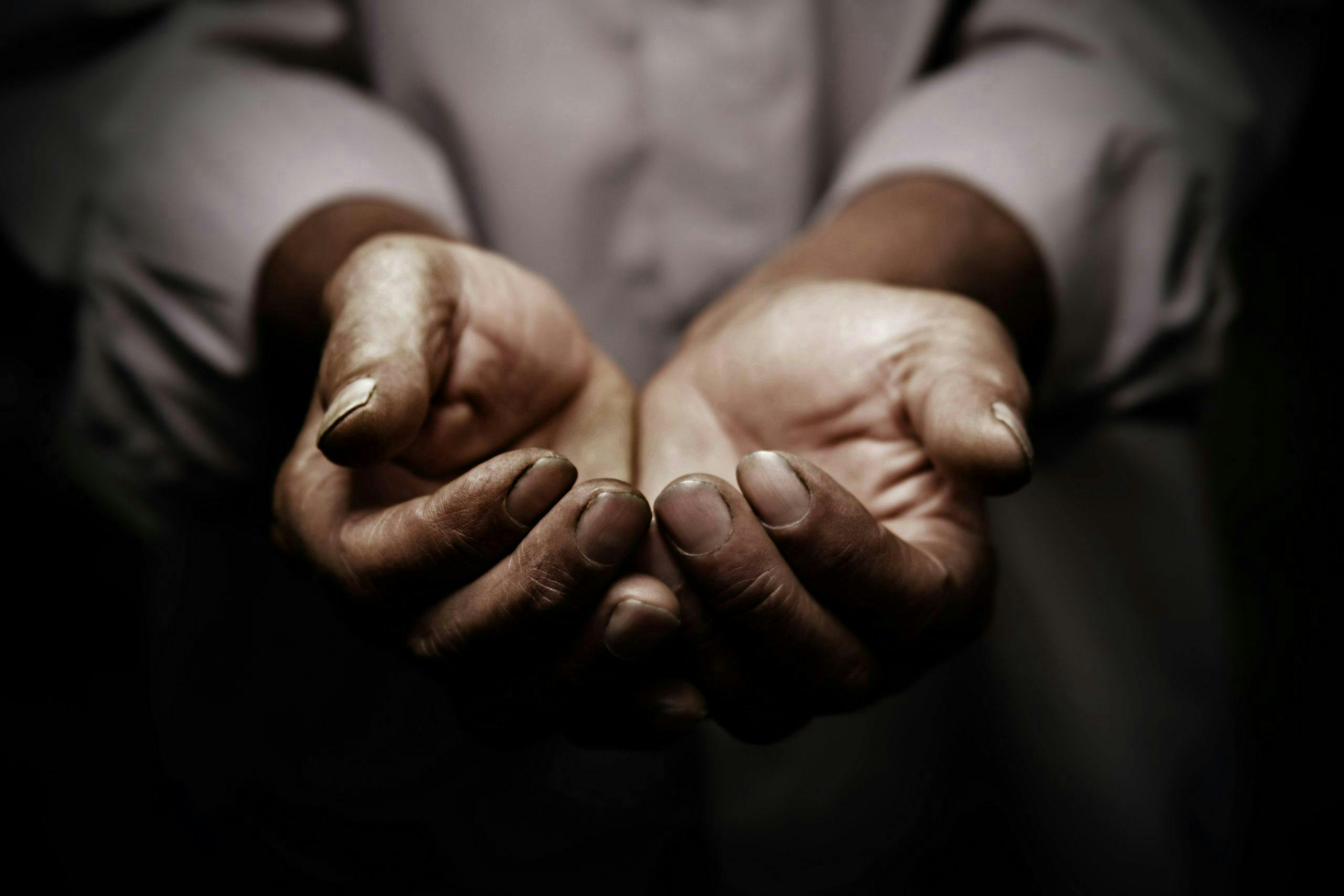 Worn hands are extended in a cupping gesture with palms facing up, set against a dark, blurred background, emphasizing the texture and appearance of the hands.