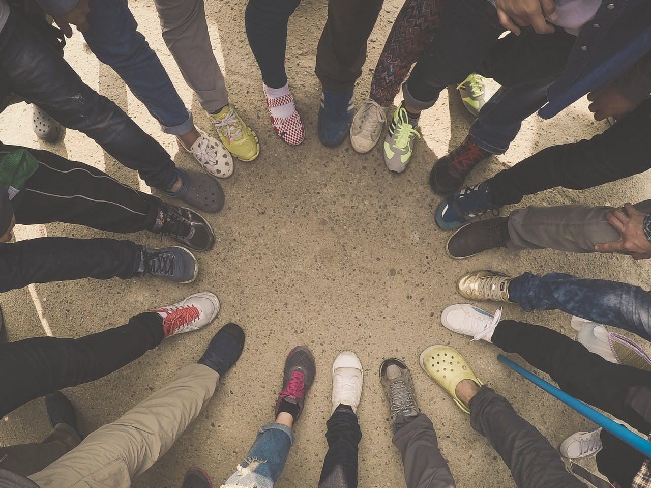 Feet in various shoes stand in a circle on a sandy ground, with visible legs suggesting a diverse group of people in a casual gathering.