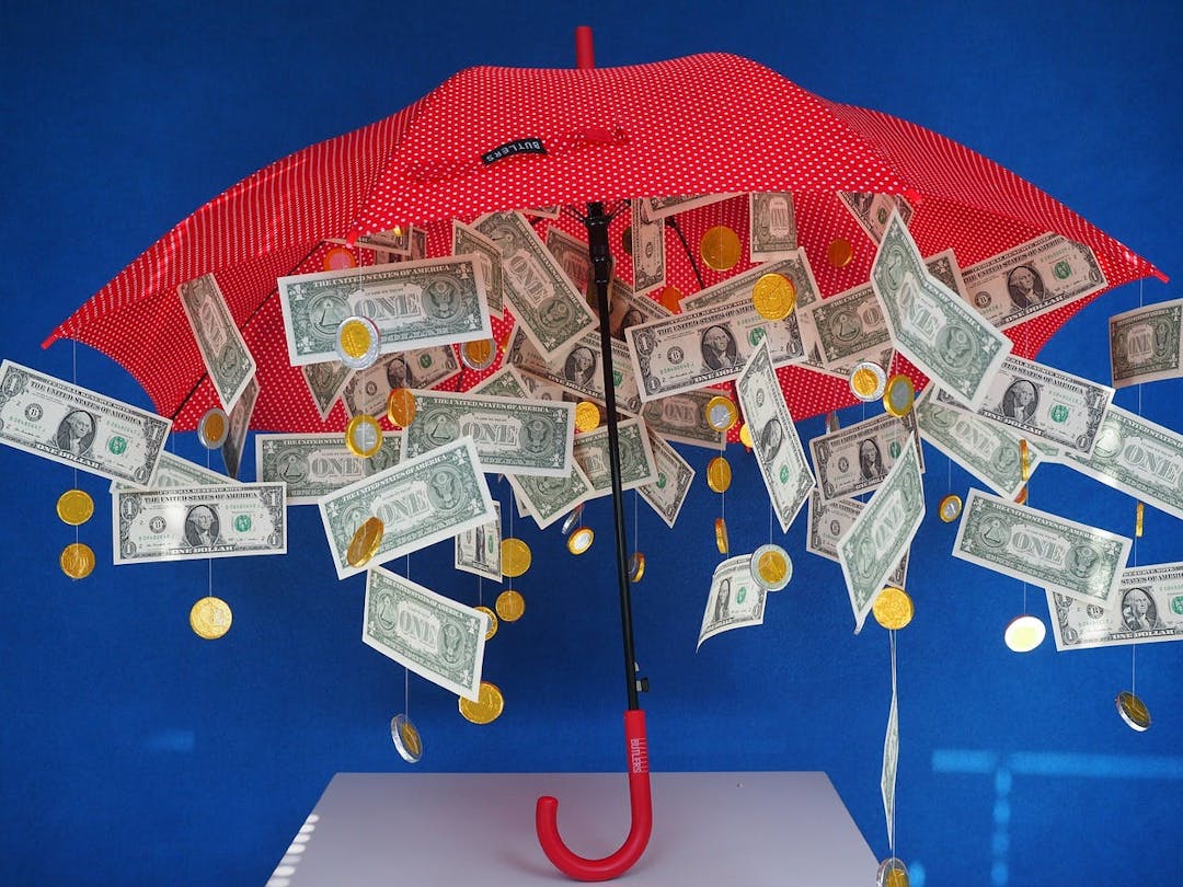 A red umbrella covered with $1 bills and gold coins is hanging upside down; the background is blue.
