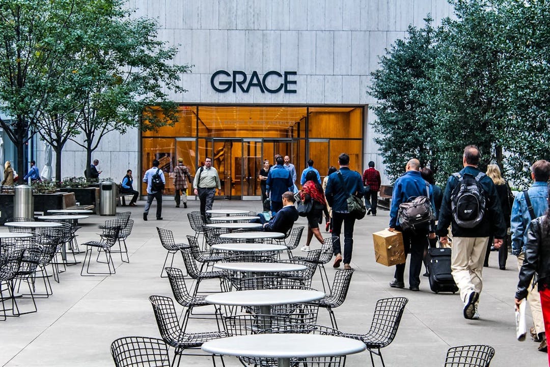 Tables and chairs sit in an outdoor plaza as people walk towards a building entrance labeled "GRACE" surrounded by trees and large windows.