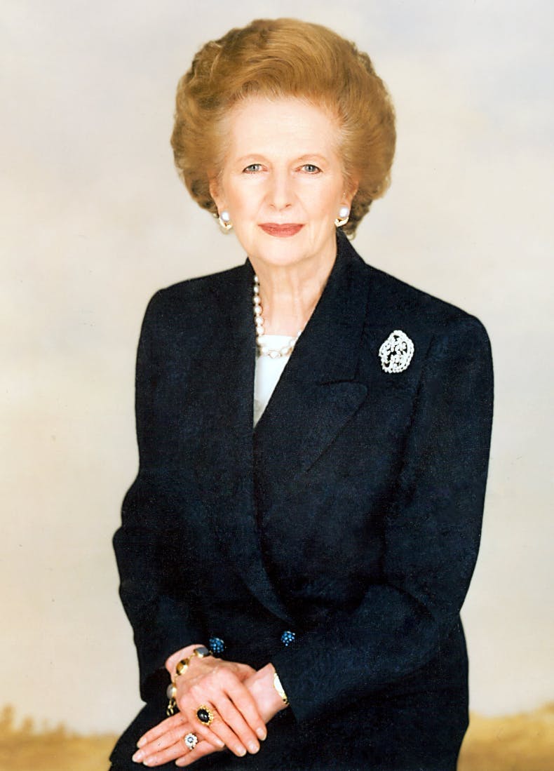 A woman dressed in a dark suit with a brooch and pearl necklace poses with clasped hands against a plain background.