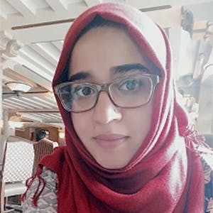 A person wearing glasses and a red hijab looks at the camera, seated indoors with a light wooden ceiling and a white lattice chair in the background.