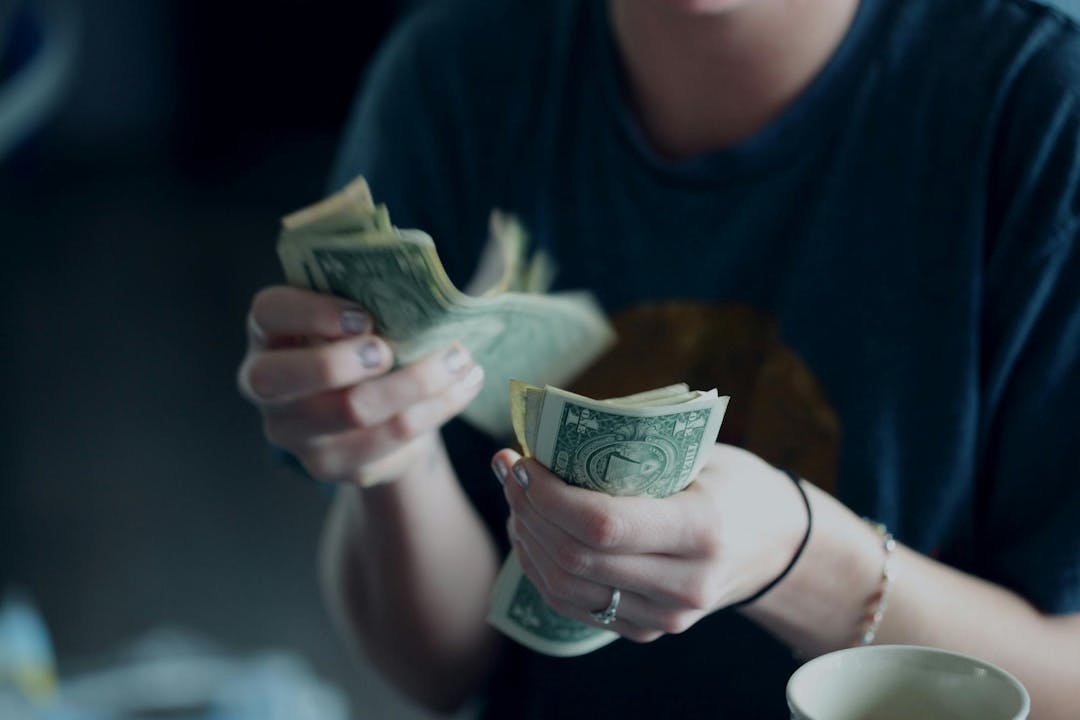 A person counts a stack of dollar bills, wearing a dark shirt with rolled-up sleeves, in an indoor environment with a white cup on the table.