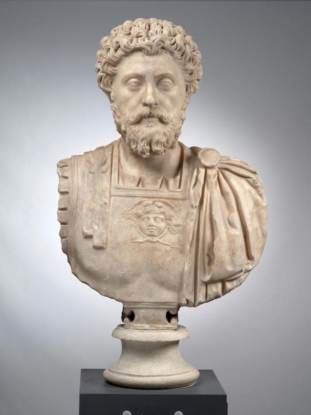 A marble bust of a bearded man in a draped garment stands on a pedestal, featuring a relief of a face on his chest, against a plain grey background.
