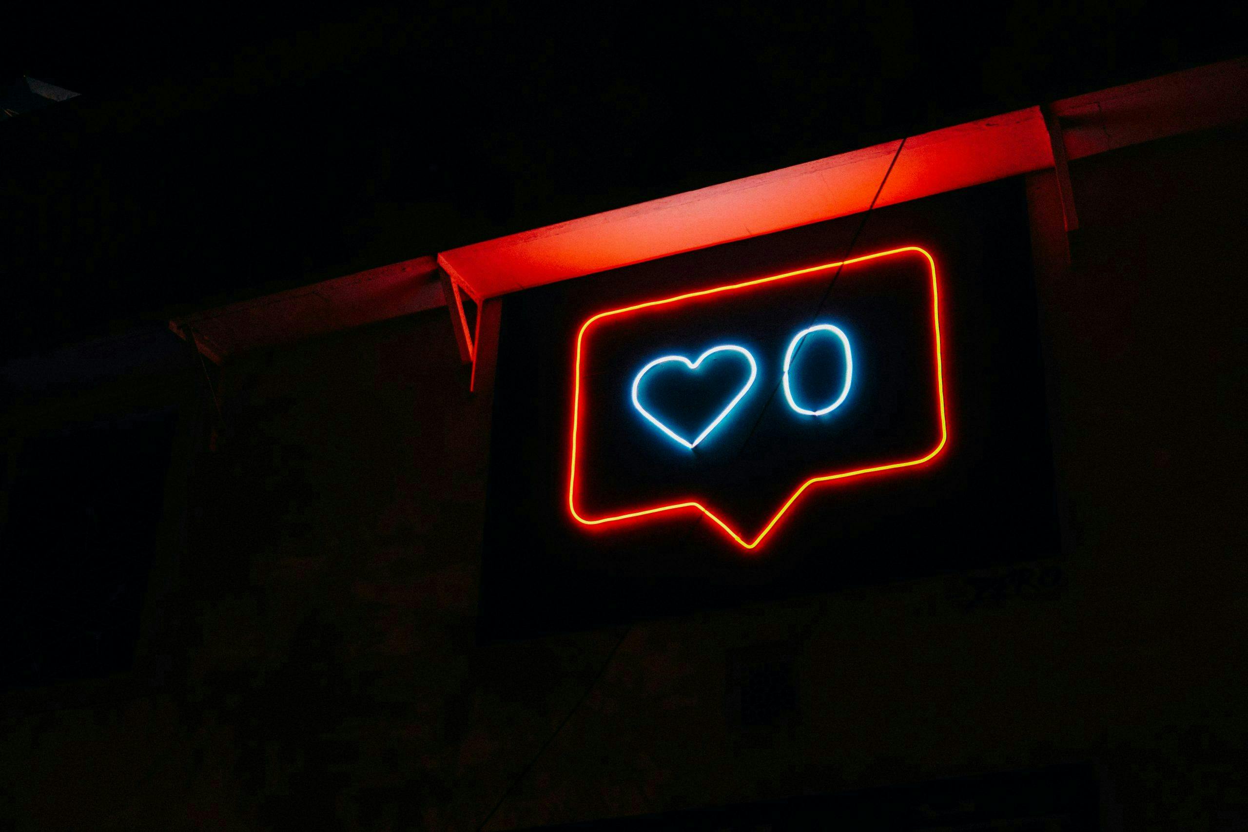 A neon sign displays a heart and the number zero inside a message bubble shape, glowing in blue and red against a dark background.