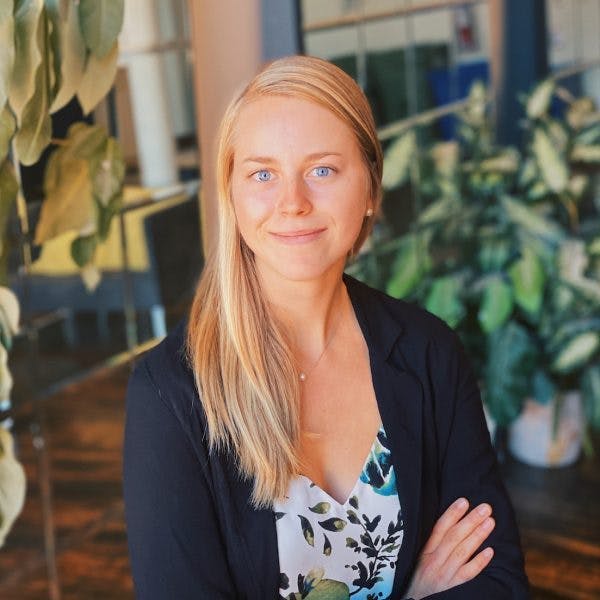 A woman with long blonde hair smiles while sitting with crossed arms, wearing a floral top and black blazer, in an office with plants and glass walls in the background.