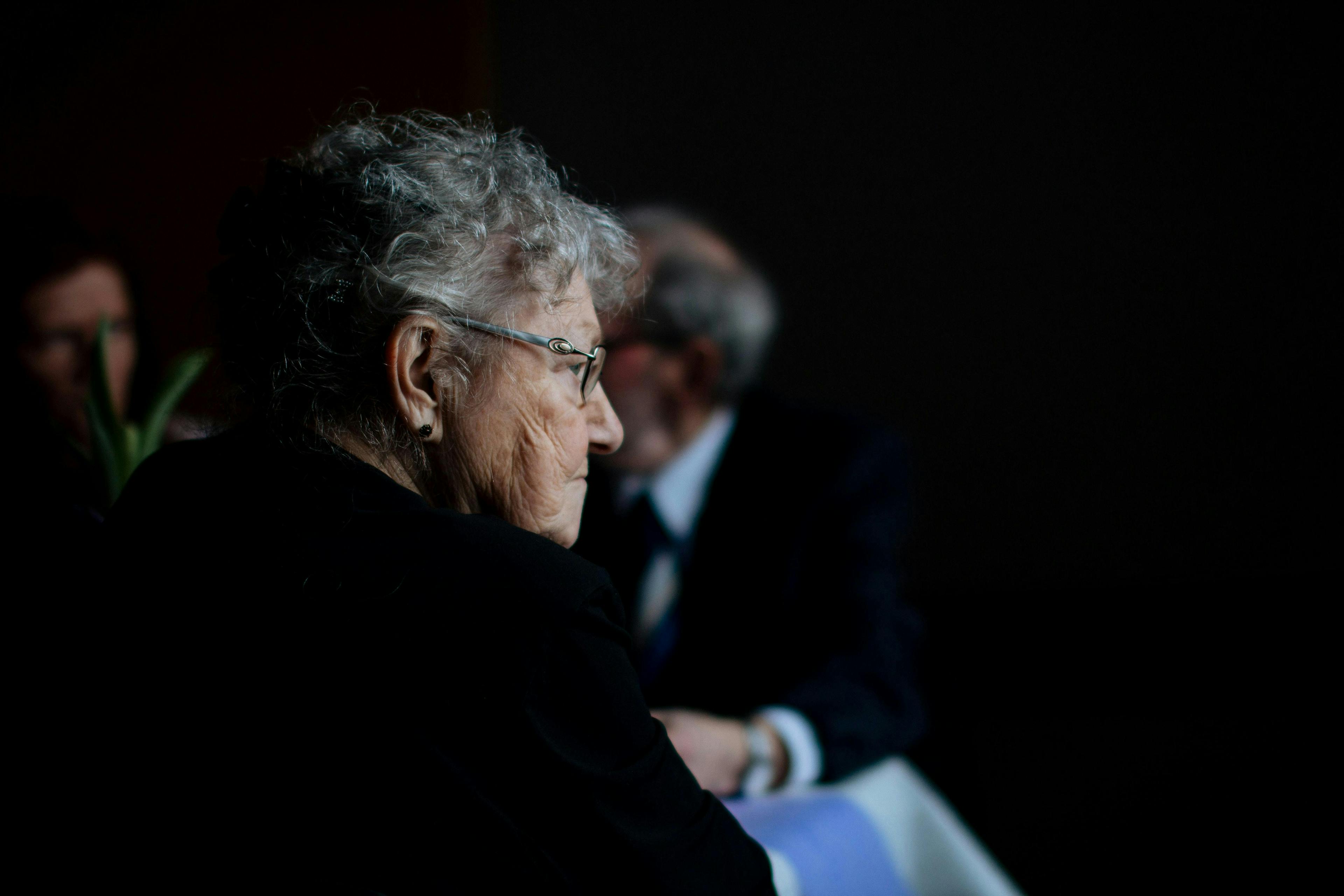 An elderly woman with gray hair and glasses sits in profile, observed in dim lighting, while another person, blurred in the background, sits at a table.