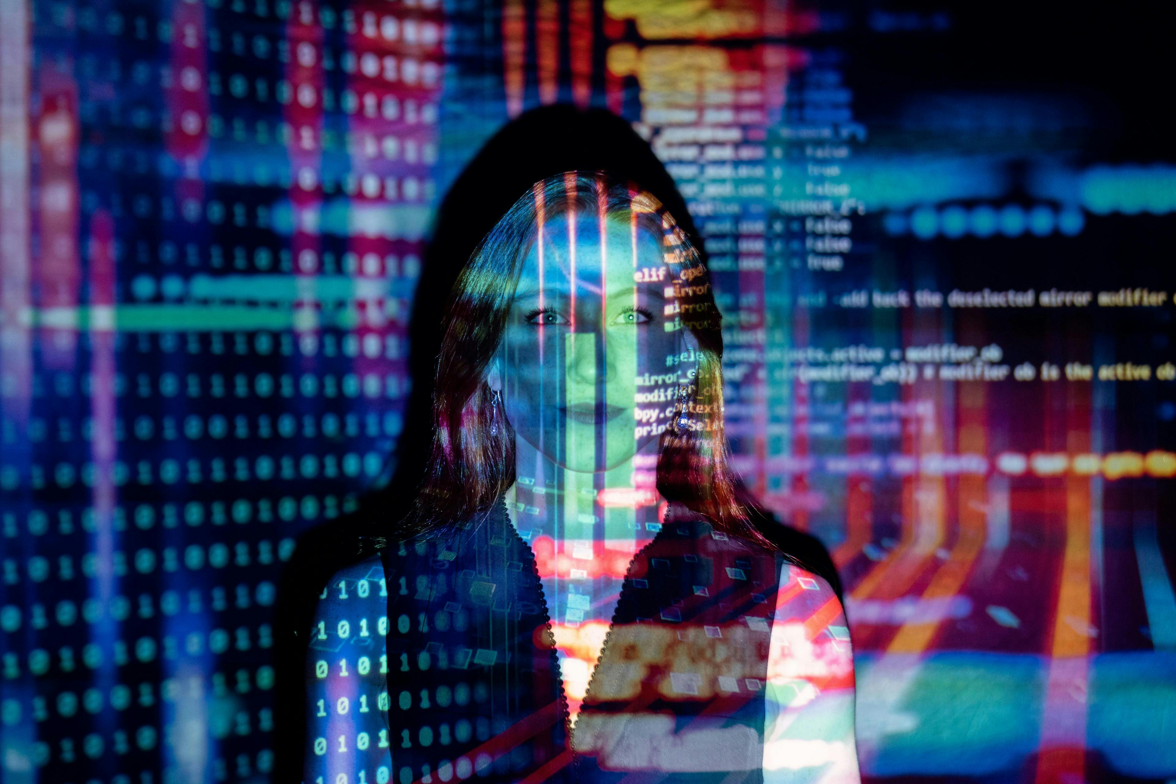 Woman standing with colorful lines of code and binary digits projected on her and the background, creating an overlay of digital imagery.