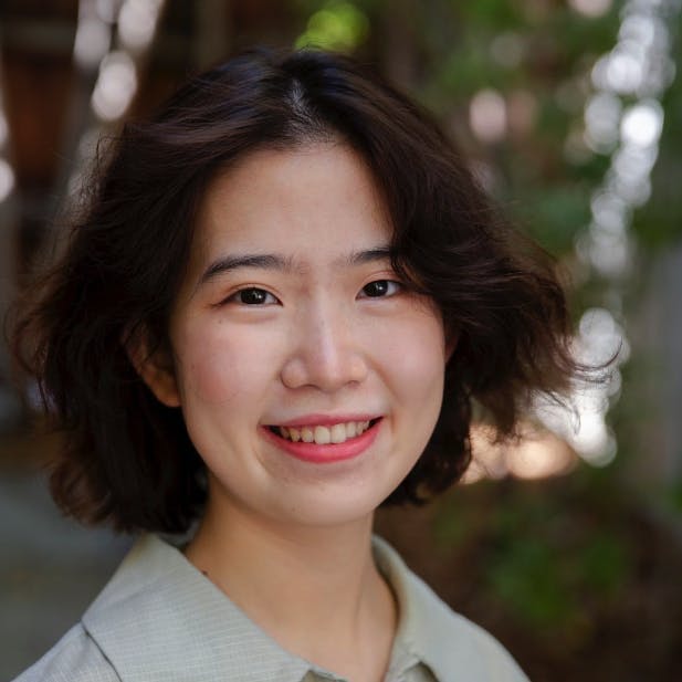 A person with short, wavy hair smiles at the camera, set against a blurred outdoor background with greenery and dappled sunlight.