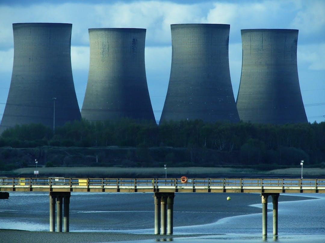 Four large cooling towers stand idle; located near a wooded area and river. A bridge with railings and pylons crosses in the foreground, with a cloudy sky above.