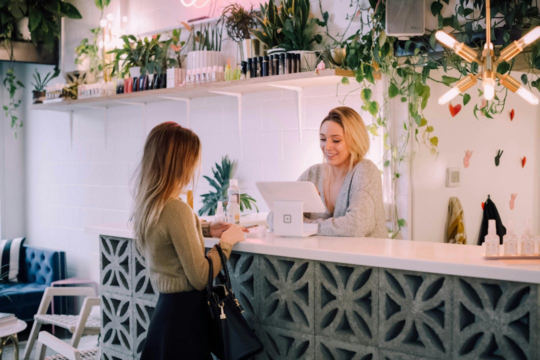 A woman stands at a counter interacting with a smiling cashier in a bright, modern store adorned with plants and shelves of products. A geometric concrete counter and a retro light fixture add to the decor.