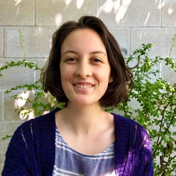 A woman is smiling, wearing a blue cardigan over a striped shirt, standing against a concrete wall with green plants and dappled sunlight in the background.