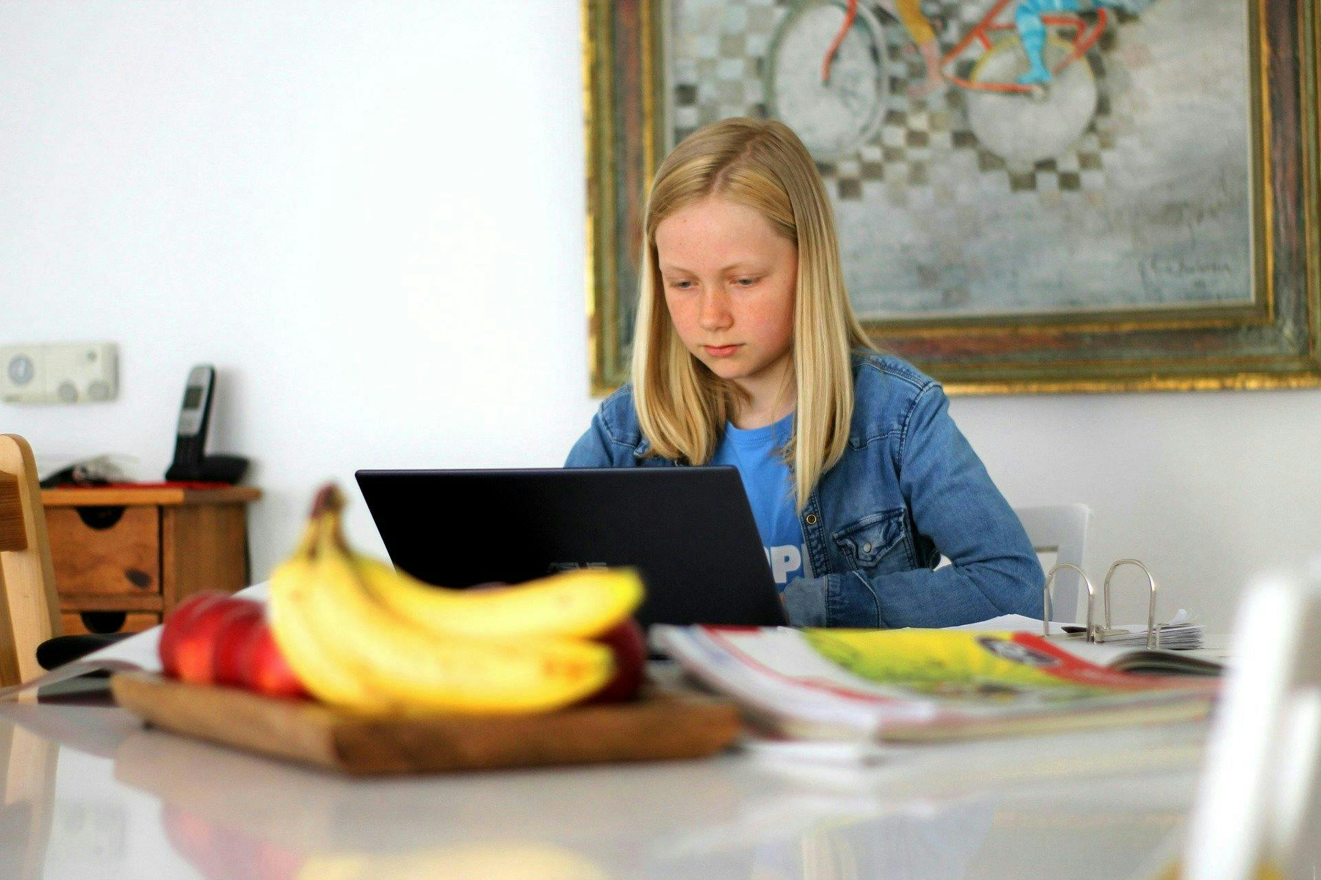 A girl works on a laptop at a white table, with bananas, a phone, and assorted papers nearby, in a room decorated with a colorful bicycle painting.