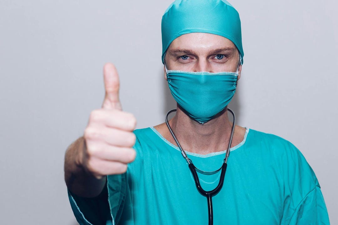 A healthcare worker in teal scrubs, cap, and mask gives a thumbs-up; a stethoscope hangs around their neck against a plain, light gray background.