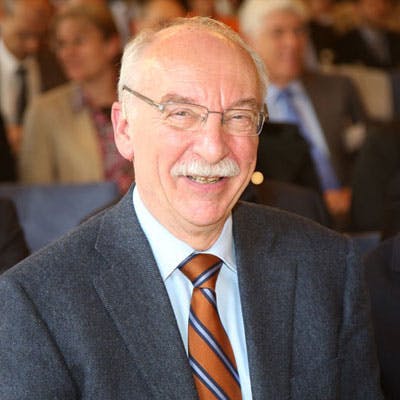 A smiling man with glasses and a mustache is sitting among a group of people in formal attire in a crowded auditorium.