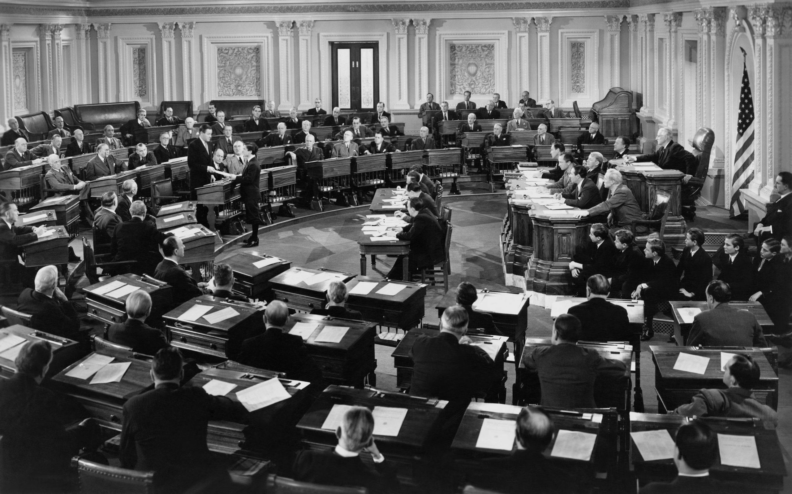 Legislators convene in a large, ornate chamber, engaged in discussions and deliberations. Wooden desks scattered with papers fill the room, and the American flag stands in the background.