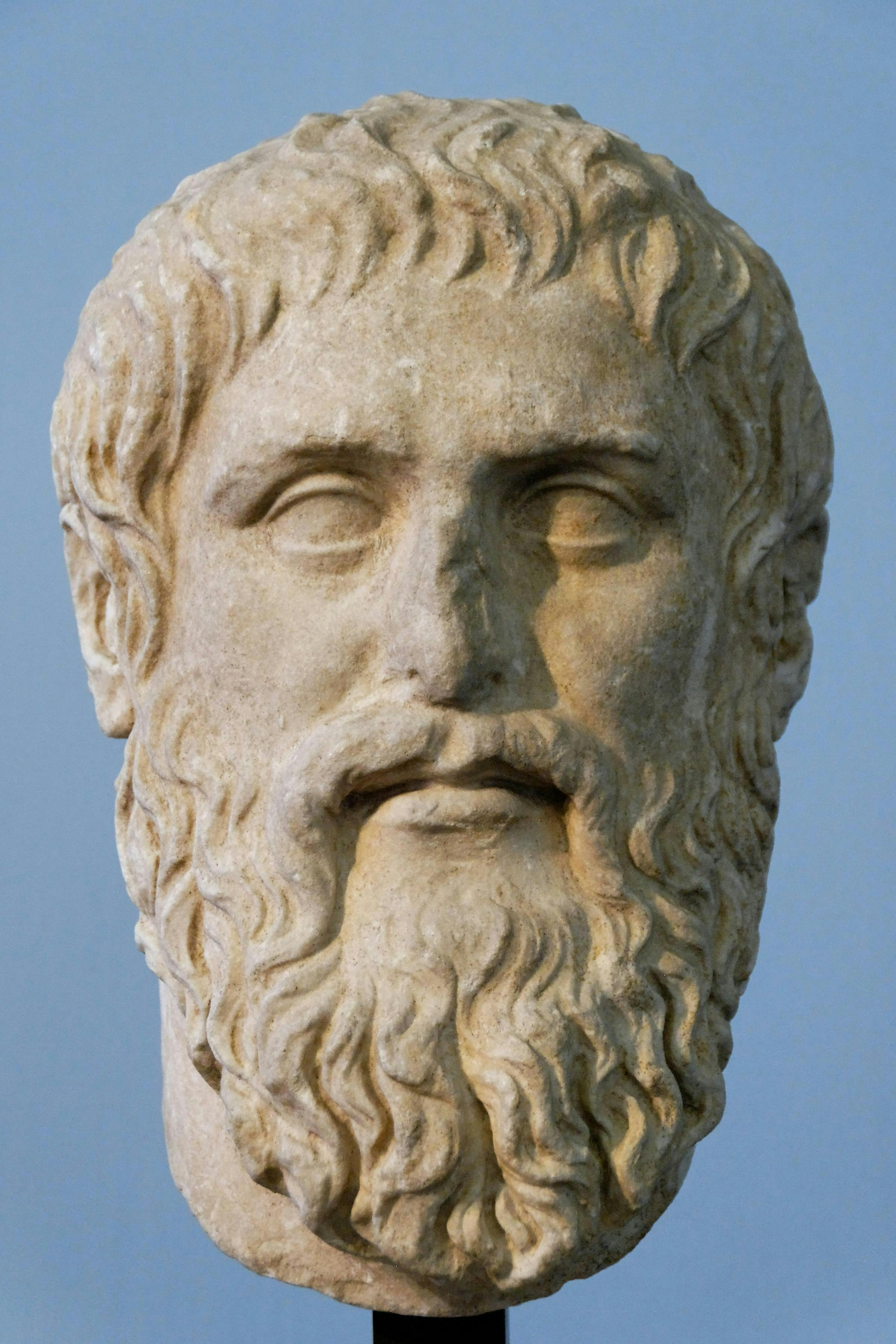 A stone bust of an ancient bearded man is displayed against a plain blue background, showcasing intricate detailing on facial features and hair.