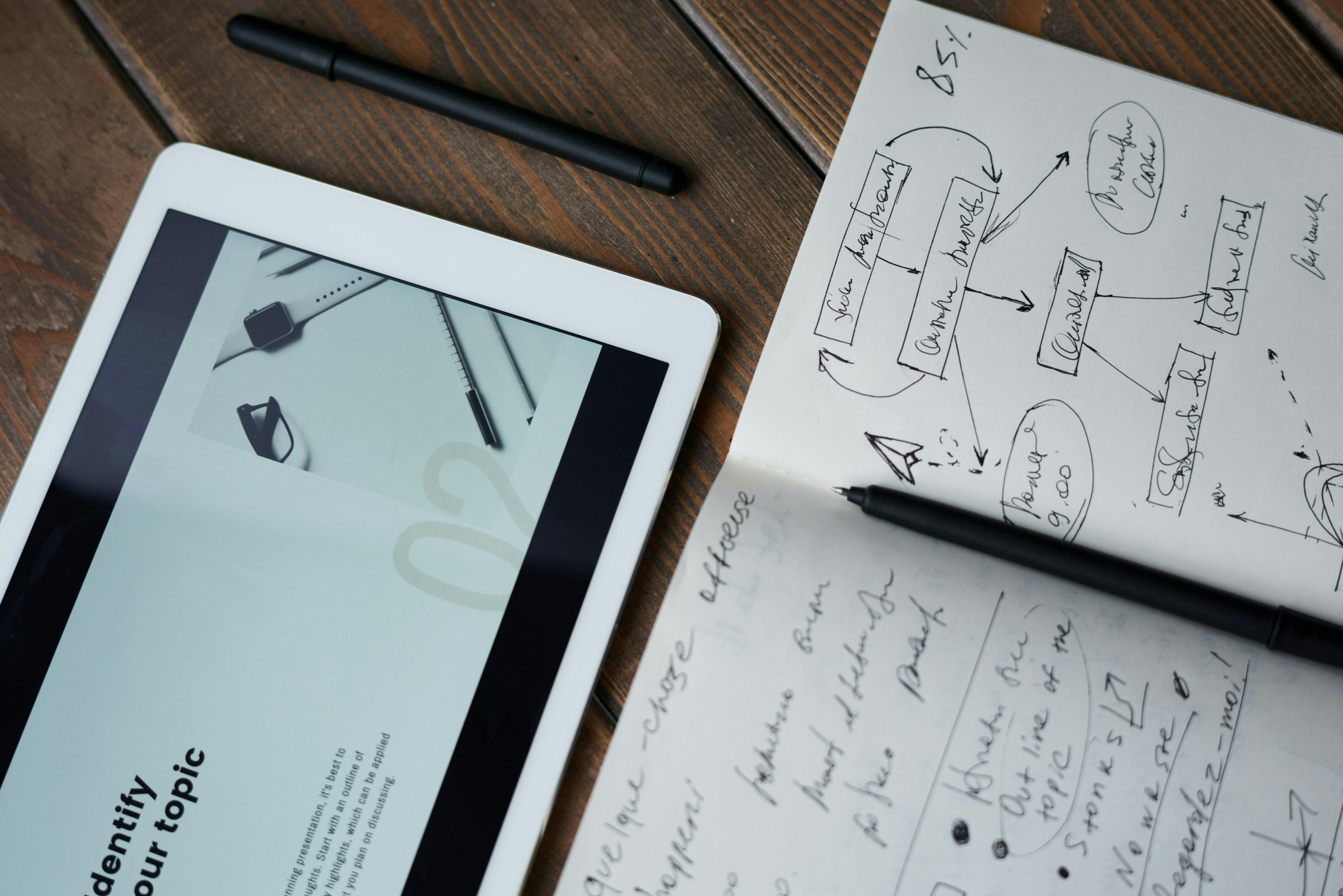 Tablet displays a presentation while a notebook with handwritten notes and diagrams lies beside it on a wooden table. Two black pens are placed near the tablet and notebook.
