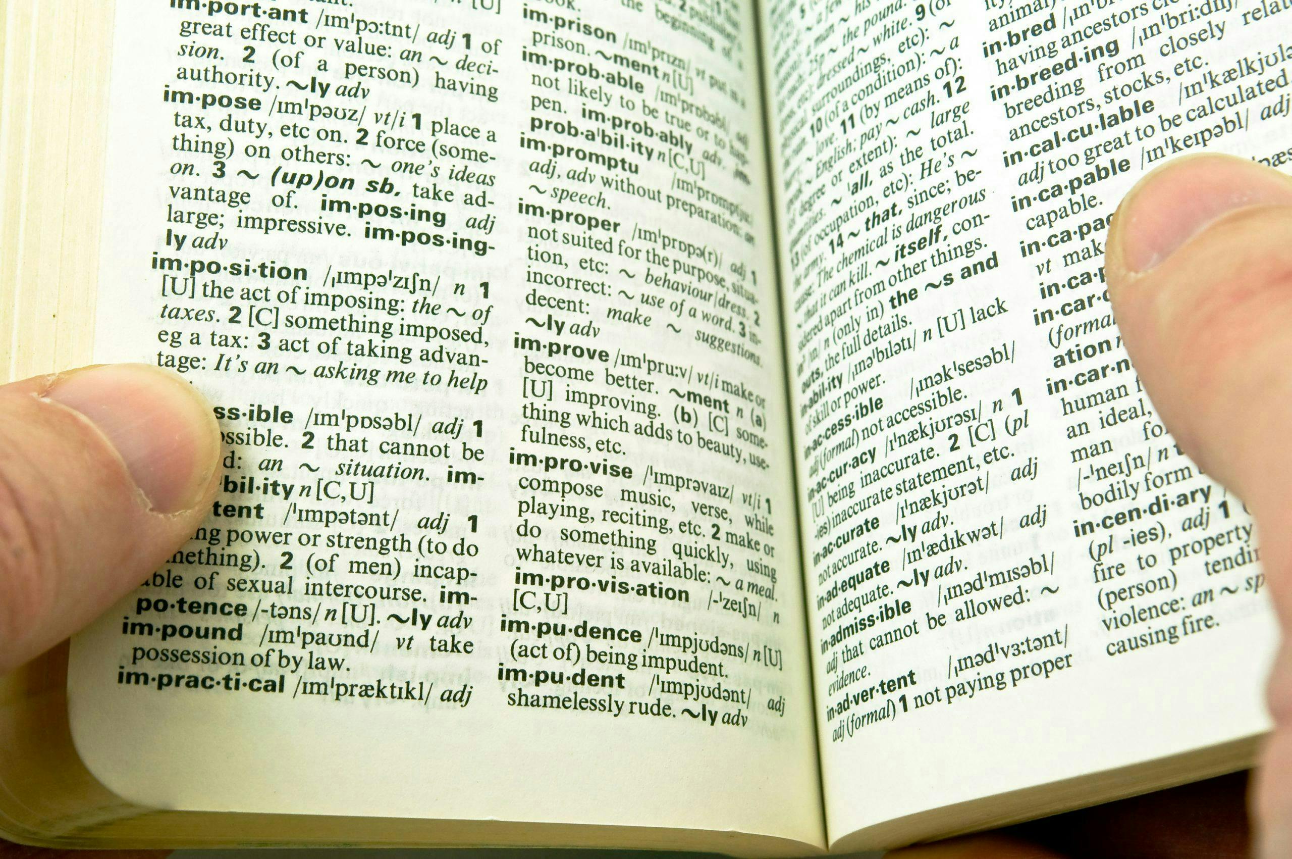A person holds open a dictionary, revealing definitions of various words on glossy pages, including "impose," "impractical," "imprison," "improve," and "improper."